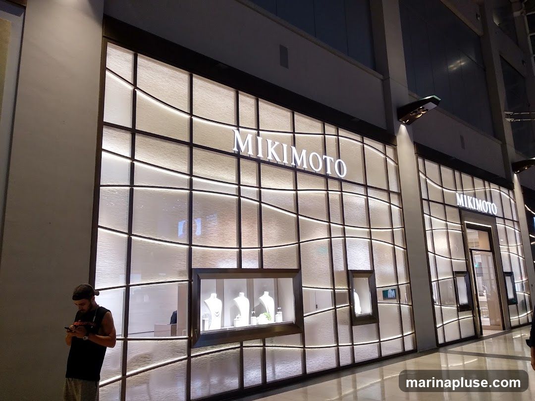 A well-lit storefront with the name MIKIMOTO displayed prominently in large white letters. The facade features a textured glass design with horizontal and curved metal accents, creating a modern and elegant aesthetic. Display windows showcase jewelry on mannequins. A person stands to the left, looking at their phone.
