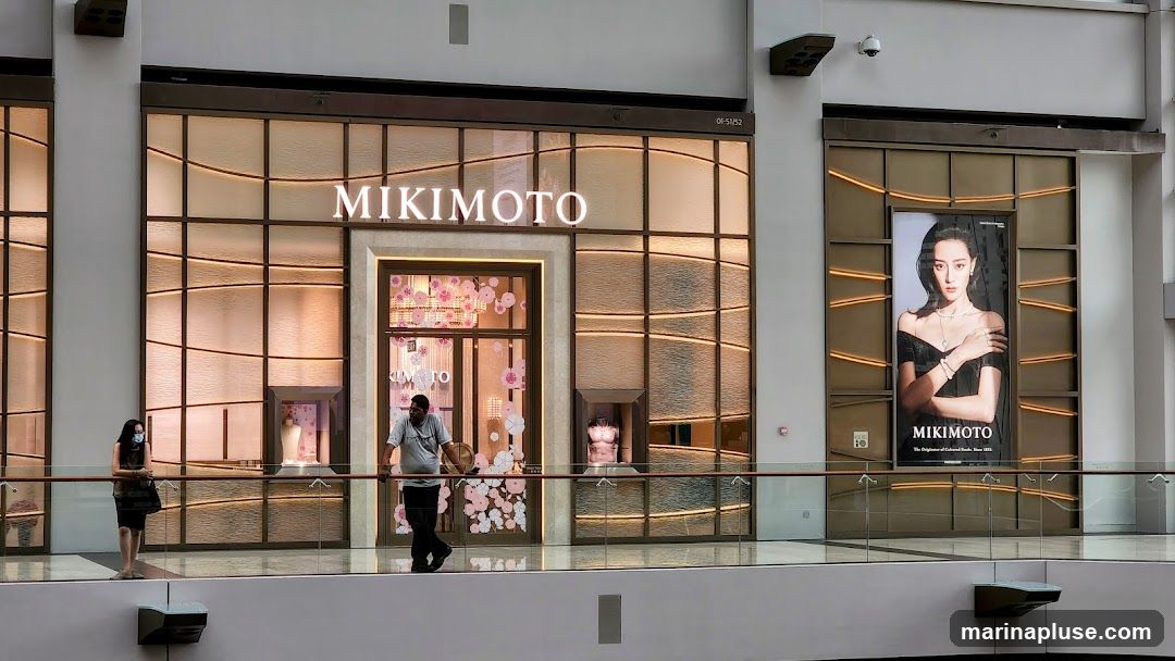 The exterior of a Mikimoto jewelry store. The storefront features a large display window with a mannequin and decorative floral elements. Above the entrance, the Mikimoto logo is prominently displayed. To the right, a large advertisement featuring a model wearing Mikimoto jewelry is visible. Two individuals are standing on a walkway in front of the store.