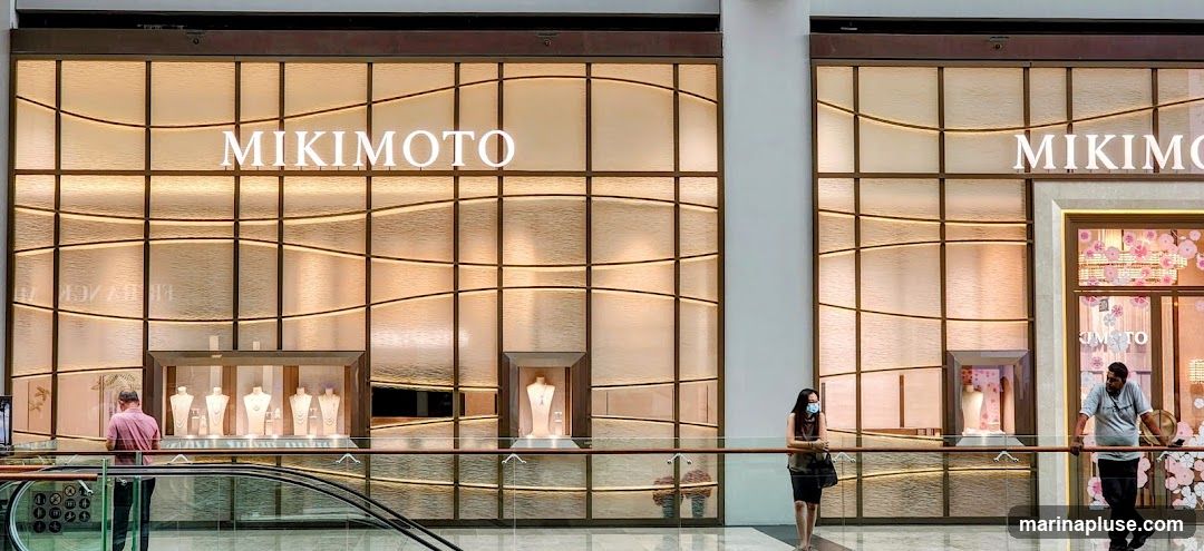 The exterior of a Mikimoto jewelry store. The storefront features large windows with a textured, backlit design. The Mikimoto logo is prominently displayed in large white letters above the windows. Inside the windows, jewelry is displayed on mannequins and stands. Two people are visible near the storefront, one looking at the displays and another standing near the entrance.