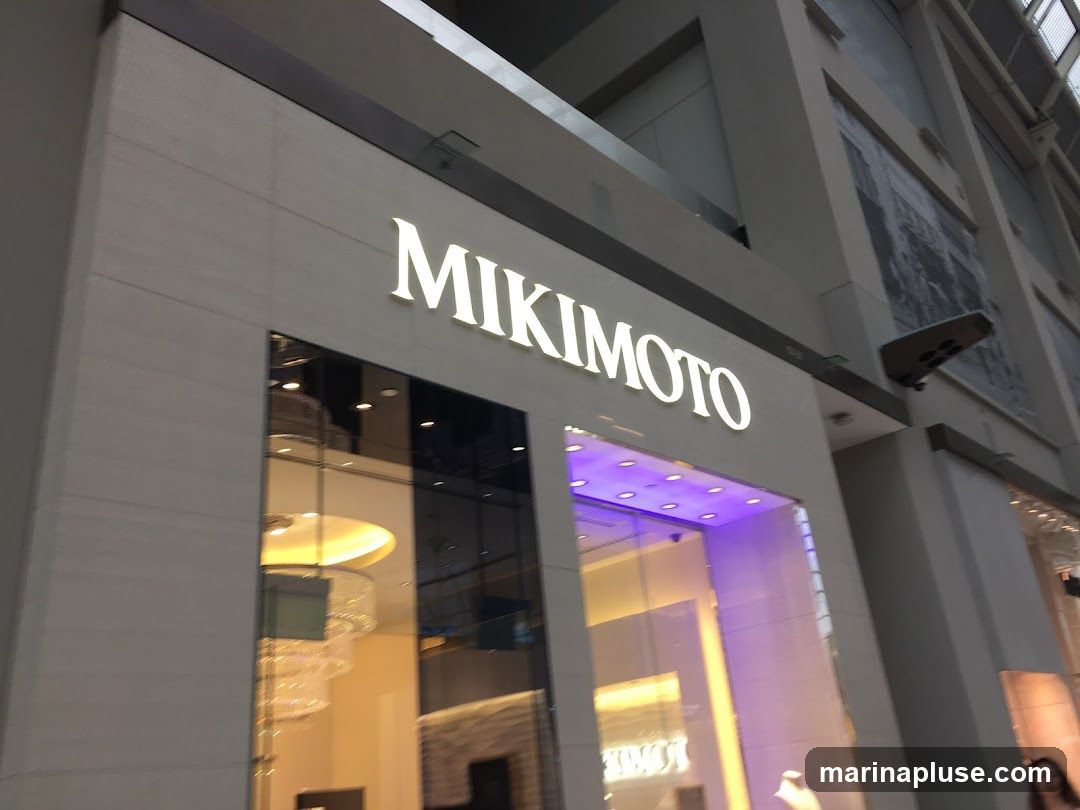 The exterior of a Mikimoto store with the brand name illuminated in large white letters above the entrance. The storefront features large glass windows reflecting the interior, which includes a chandelier and display cases. The facade is made of light-colored stone panels.