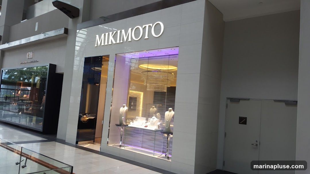 The exterior of a Mikimoto jewelry store. The store has a large display window showcasing necklaces on mannequins and a chandelier inside. The Mikimoto logo is visible above the window. To the left, another store with the logo "CH" is visible.