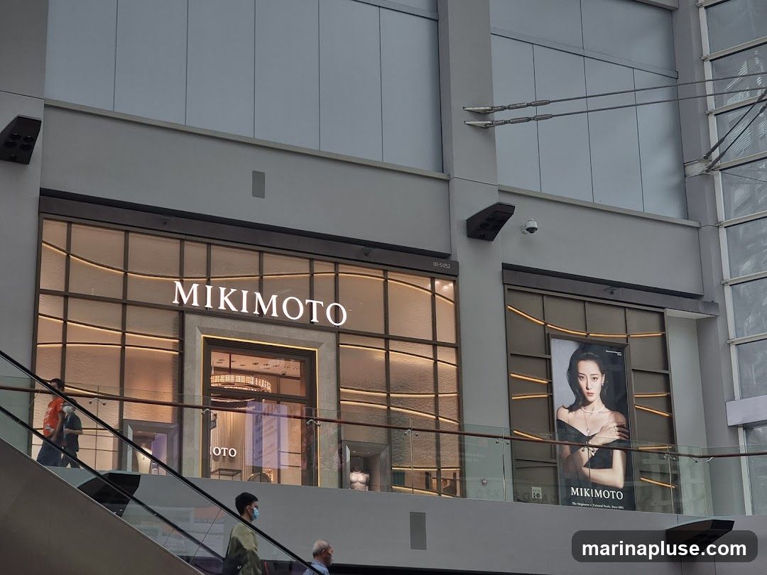 The exterior of a Mikimoto store is visible, with the brand name illuminated above the entrance. The store features a large display window with a grid pattern and warm interior lighting. To the right of the entrance, a large advertisement featuring a woman wearing jewelry is displayed. People are seen on an escalator in the foreground.