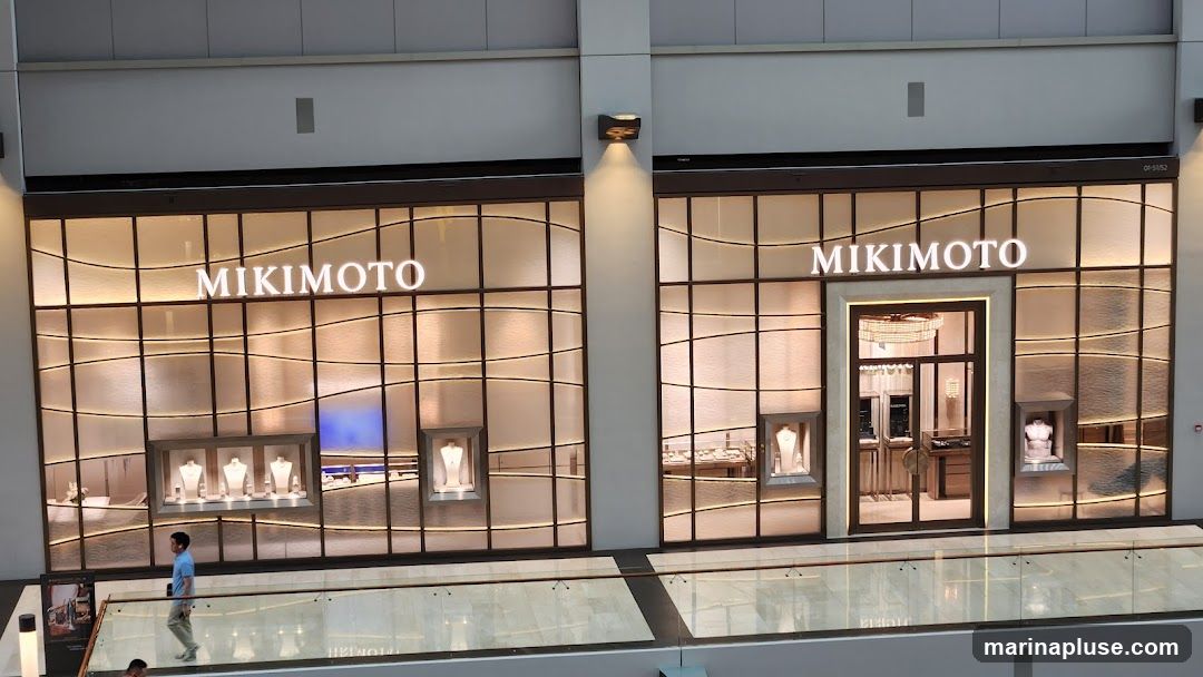 The exterior of a Mikimoto jewelry store. The storefront features large glass windows with a wavy pattern and illuminated displays showcasing necklaces and busts. The Mikimoto logo is prominently displayed in large white letters above the windows. A central entrance with double glass doors is visible, leading into the store where more displays and lighting can be seen. A person is walking in front of the store on a walkway.