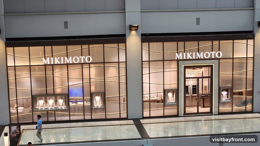 The exterior of a Mikimoto jewelry store. The storefront features large glass windows with a wavy pattern and illuminated displays showcasing necklaces and busts. The Mikimoto logo is prominently displayed in large white letters above the windows. A central entrance with double glass doors is visible, leading into the store where more displays and lighting can be seen. A person is walking in front of the store on a walkway.