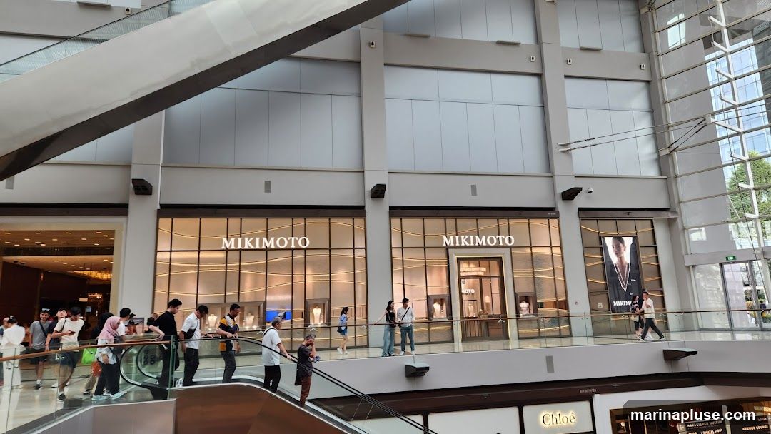 The exterior of a Mikimoto store is visible, with large windows displaying jewelry. The store's name is prominently displayed in large white letters above the windows. People are seen walking on a walkway in front of the store and on escalators below.