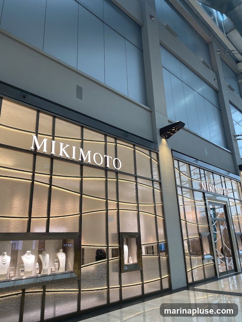 The exterior of a Mikimoto jewelry store. The storefront features large windows displaying jewelry on mannequins and stands. The store's name, MIKIMOTO, is prominently displayed in illuminated lettering above the entrance and on the facade. The building has a modern design with textured glass panels and warm interior lighting visible through the windows.