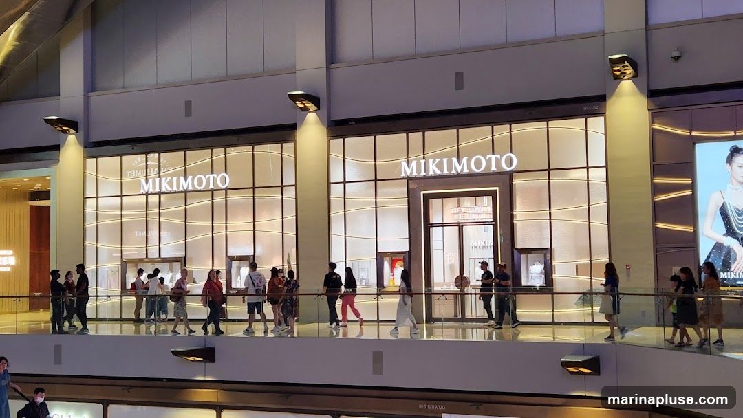 A wide shot of the Mikimoto store facade in a mall. The store has a large glass front with a prominent "MIKIMOTO" sign above the entrance. Several people are walking along the railing in front of the store. To the right of the store is a large advertisement featuring a woman wearing jewelry.