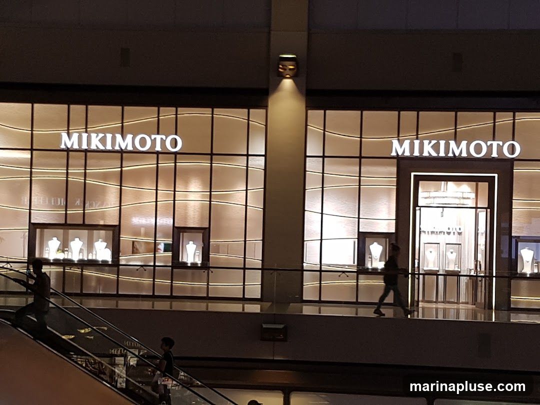 The exterior of a Mikimoto jewelry store is shown at night. The store has a large glass facade with the Mikimoto logo displayed prominently in illuminated white letters above the entrance. Inside, jewelry displays are visible, showcasing necklaces and other items on mannequins. The store is lit from within, creating a warm glow. People are seen walking past the store and on an escalator in the foreground.