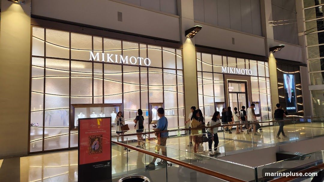 The exterior of a Mikimoto jewelry store in a modern shopping mall. The storefront features large glass windows displaying jewelry, with the Mikimoto logo prominently displayed above the entrance. Several people are seen browsing outside the store.