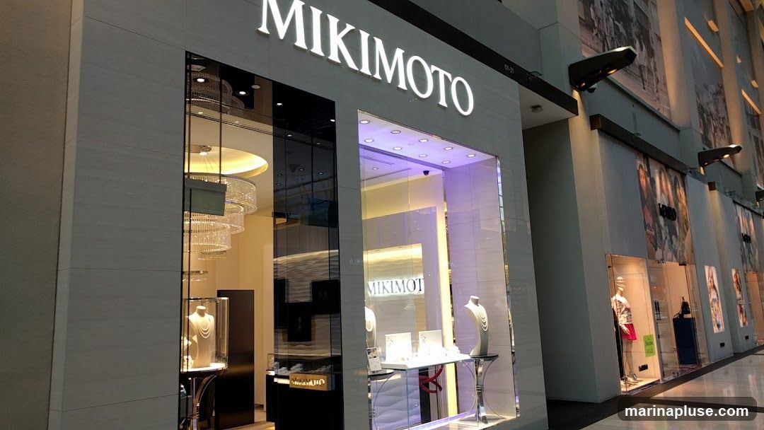 The exterior of a Mikimoto jewelry store. The store has a large, illuminated sign that reads "MIKIMOTO" above a glass storefront. Inside, jewelry is displayed on mannequins and stands. The store features elegant lighting and modern design.