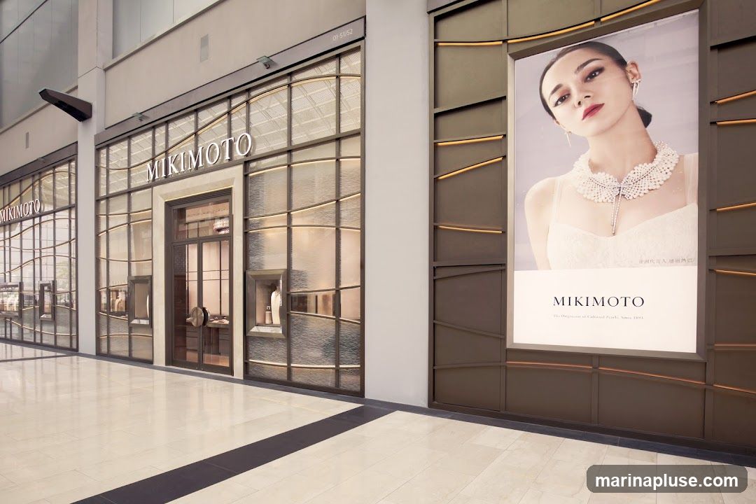 The exterior of a Mikimoto jewelry store. The storefront features large glass windows with decorative metal framing, displaying jewelry inside. The Mikimoto logo is prominently displayed above the entrance. To the right of the entrance is a large advertisement featuring a woman wearing Mikimoto pearls.