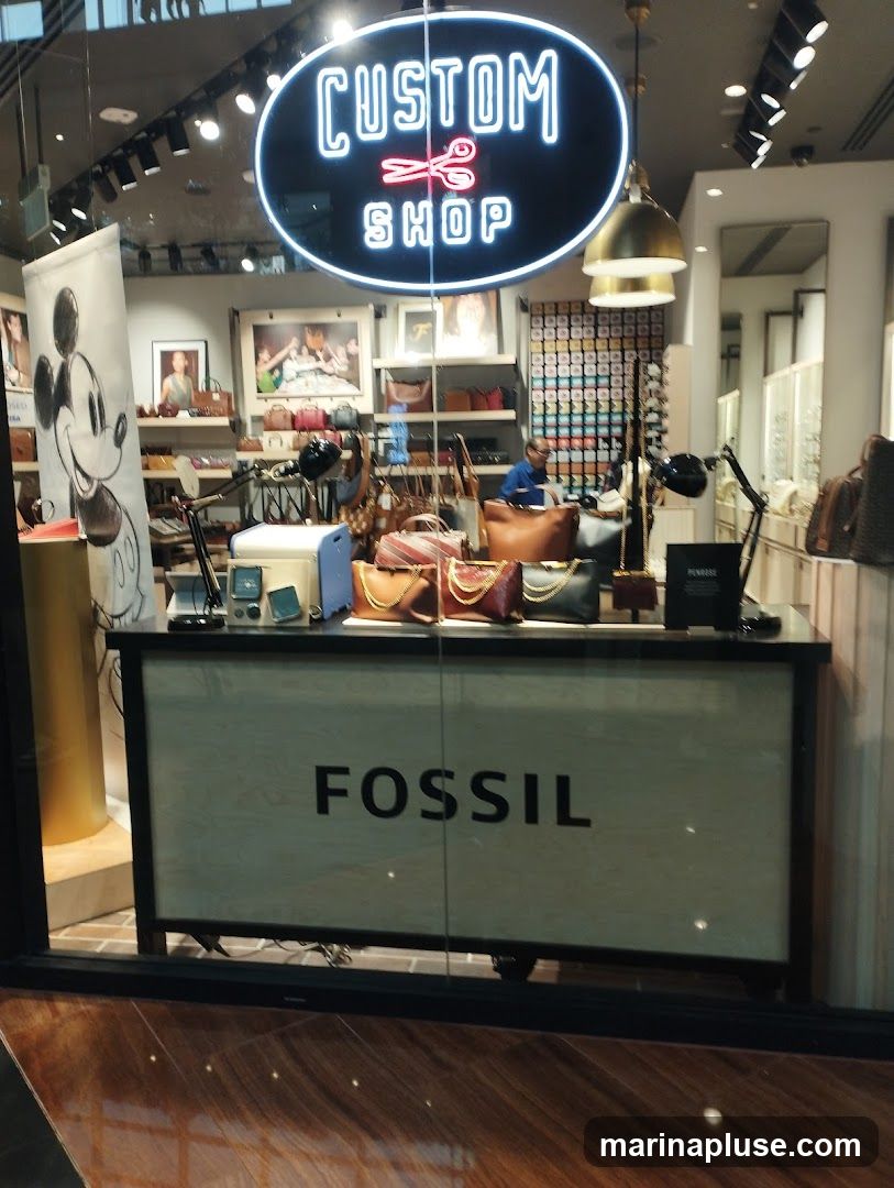 A display window for a Fossil store features a neon sign that reads "CUSTOM SHOP" with scissors in the middle. Below the sign, a large banner displays the word "FOSSIL". The display showcases various handbags and accessories. A large cutout of Mickey Mouse is visible on the left side of the window.