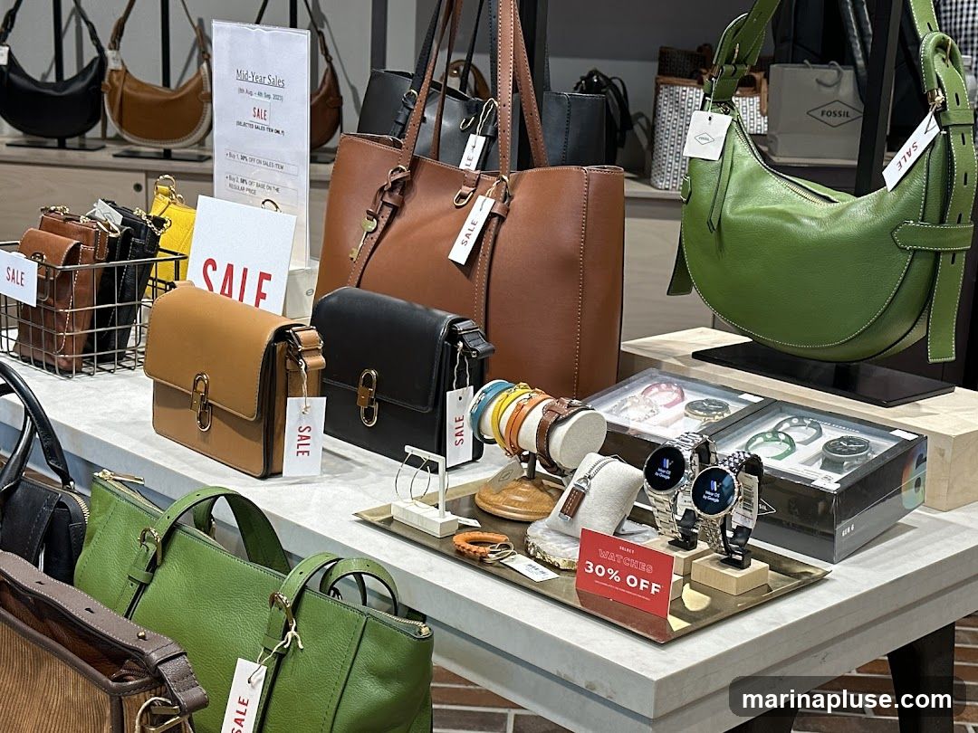 A display of Fossil handbags and watches. Several handbags in various colors including brown, black, and green are showcased. In the foreground, a display of watches is visible, with a sign indicating "WATCHES 30% OFF". A "Mid-Year Sales" sign is also present.
