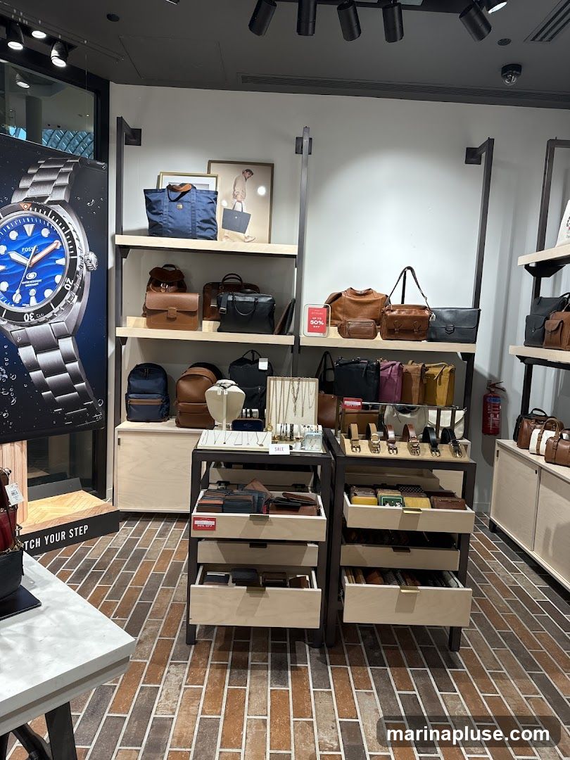 A retail store display featuring Fossil brand bags, wallets, belts, and watches. The store has a modern aesthetic with light wood shelving and brick-patterned flooring. A large advertisement for a Fossil watch is visible on the left. A "SALE 50% OFF" sign is displayed on a shelf with handbags.