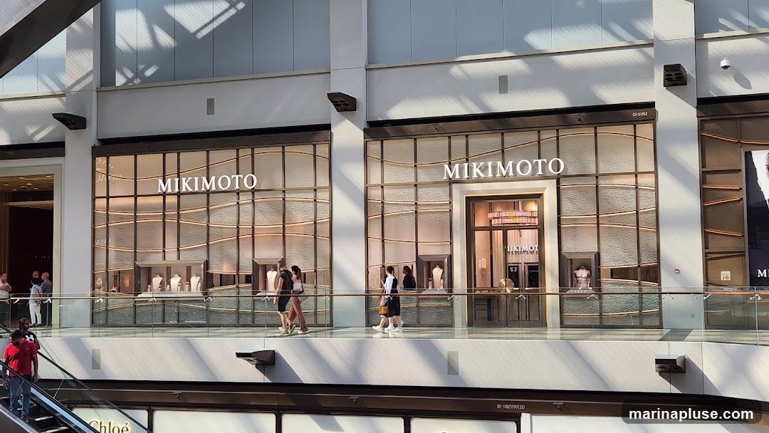 The exterior of a Mikimoto jewelry store, featuring large display windows with the brand name prominently displayed above. The windows showcase jewelry displays and mannequins. People are seen walking past the storefront on a walkway.