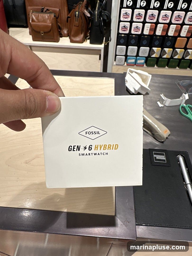 A hand holds a white box with the Fossil logo and text that reads "GEN 6 HYBRID SMARTWATCH". In the background, shelves display brown leather bags and a wall is covered with small square items in a grid pattern.