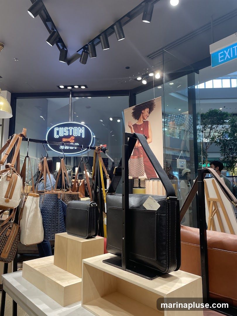 A retail display featuring various bags, including leather handbags and crossbody bags, arranged on wooden stands and hanging racks. A neon sign that reads "CUSTOM SHOP" with scissors is visible in the background. A poster of a woman with a handbag is also displayed. An "EXIT" sign is visible above the display.