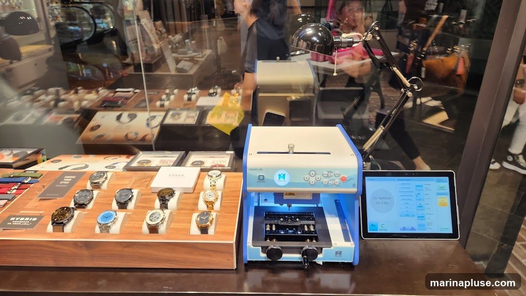 A display of Fossil watches is shown on a wooden stand. To the right of the watches is a blue and white engraving machine labeled 'MAGIC-25' with a tablet screen next to it. The tablet displays a user interface with various options and settings. The background shows reflections of people and escalators, suggesting an indoor shopping environment.
