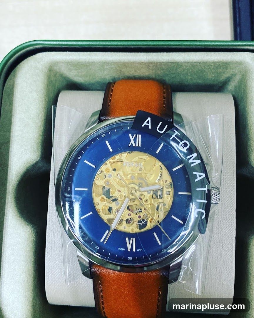 A close-up shot of a Fossil automatic watch with a blue dial and a brown leather strap, presented in its original packaging. The watch features an open-heart design, revealing the intricate mechanical movement. The dial has Roman numerals at the 12 o'clock position and Arabic numerals around the outer edge. The word "AUTOMATIC" is printed on a black tag attached to the watch.