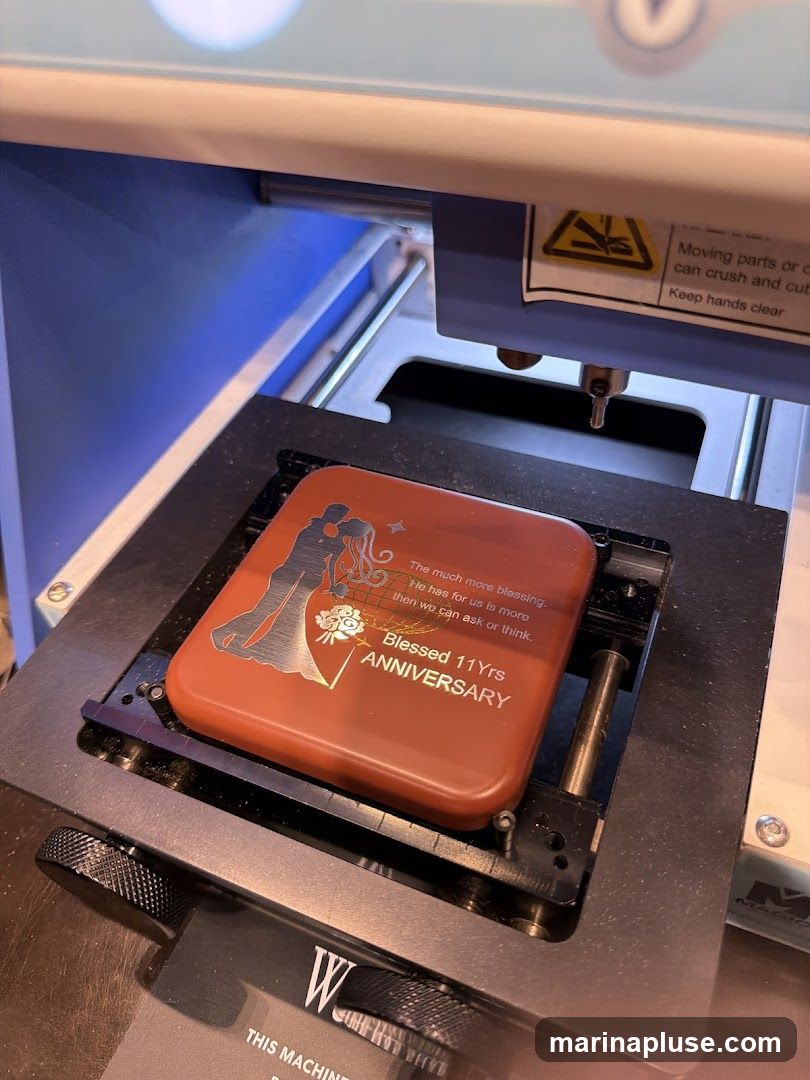 A close-up view of a laser engraving machine with a red rectangular object placed on its platform. The object has an engraved design featuring a couple in silhouette, flowers, and text that reads "The much more blessing. He has for us is more then we can ask or think. Blessed 11Yrs ANNIVERSARY." Warning labels are visible on the machine, indicating potential hazards.