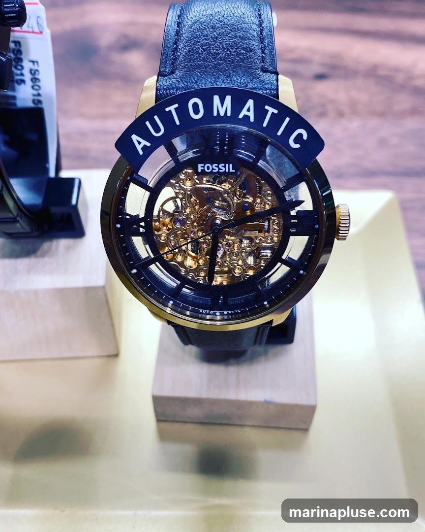 A close-up shot of a Fossil automatic watch with a black leather strap and a gold-toned skeleton dial. The watch is displayed on a wooden stand. A black curved tag above the watch face reads "AUTOMATIC". Another tag with "FS6015" is visible on the left.
