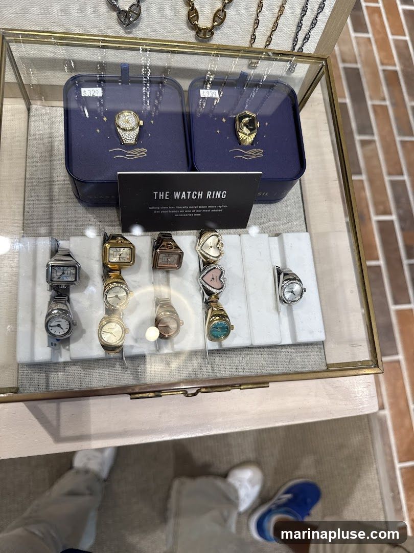 A glass display case filled with various watches and rings. Two prominent watch rings are showcased in blue boxes with prices $329 and $239. Below, a row of smaller watches are displayed on a white marble stand. The text on a card reads "THE WATCH RING. Telling time has literally never been more stylish. Set your hands on one of our most adored accessories now."