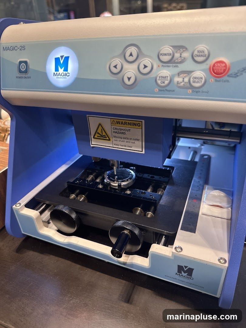 A close-up view of a MAGIC-2S engraving machine. The machine has a blue and white casing with various buttons and controls. A warning label for crush/cut hazard is visible. The machine appears to be in operation, with a small object secured in the engraving area.