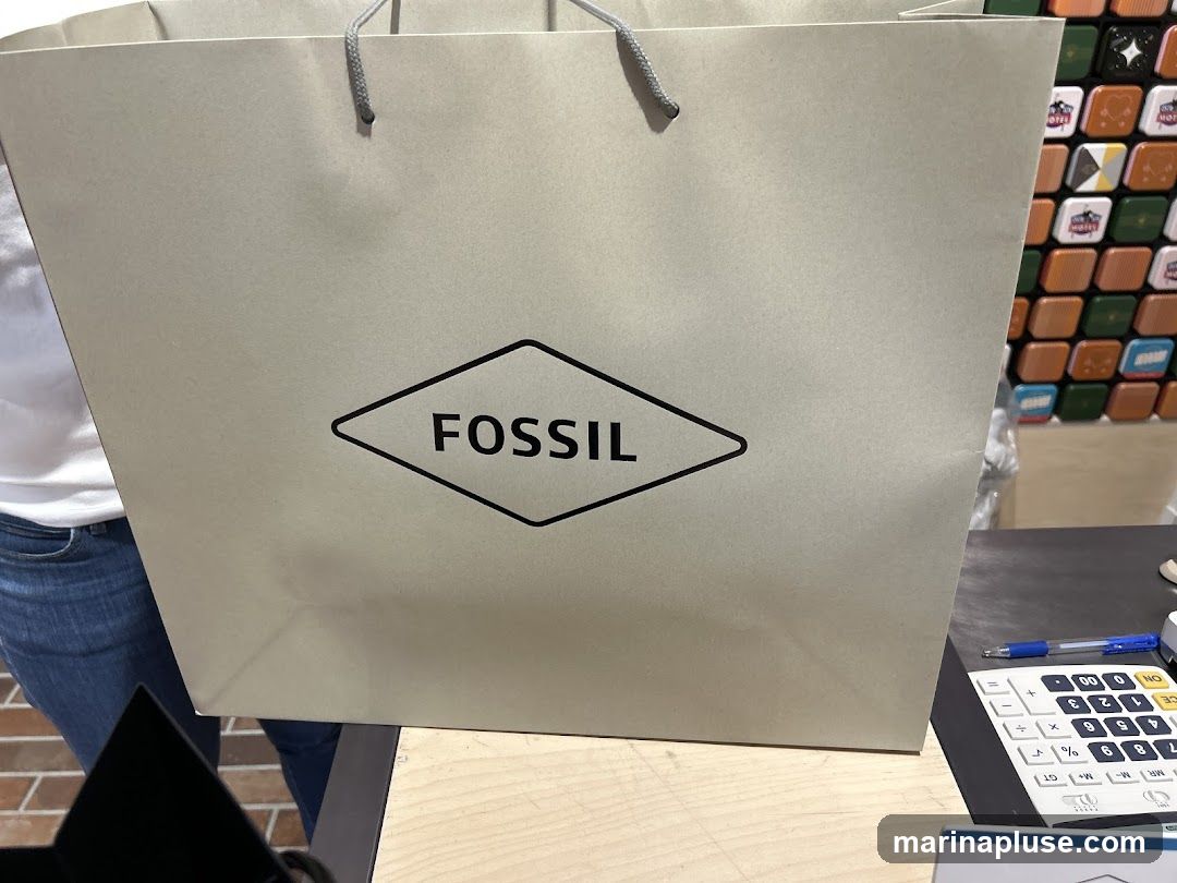 A light beige shopping bag with the Fossil logo printed in black on the front. The logo is a diamond shape with the word "FOSSIL" inside. The bag has gray rope handles. In the background, there is a wall with colorful square items and a calculator on a counter.