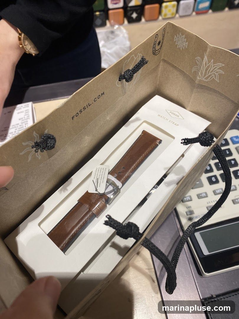 A brown leather watch strap is displayed inside a Fossil branded shopping bag. The bag has the Fossil.com logo printed on the inside. A calculator is visible to the right of the bag. In the background, there are shelves with various items, possibly watch boxes.