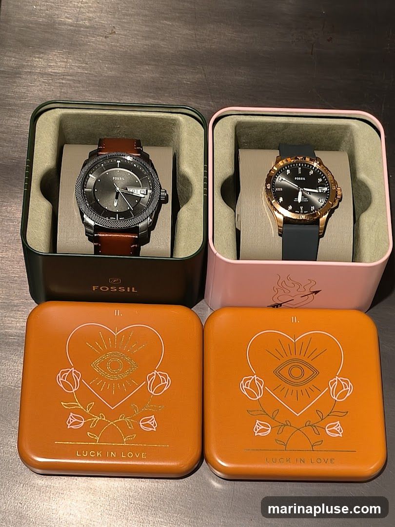 Two Fossil watches are displayed in their respective boxes. The watch on the left has a dark gray face with a brown leather strap and is in a dark green box. The watch on the right has a rose gold case, a black face, and a dark gray silicone strap, and is in a pink box. Below the watch boxes are two orange tins with a "Luck in Love" design featuring a heart with an eye and roses.