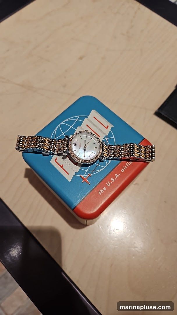 A Fossil watch with a two-tone metal band and a white face with Roman numerals is resting on a blue and red tin. The tin has a globe graphic and text that reads "the U.S.A. airli".