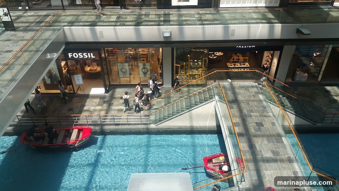 An aerial view of an indoor shopping mall with a canal running through it. Several shops are visible, including a Fossil store. People are walking on the elevated walkways and stairs, and some are in boats on the canal.