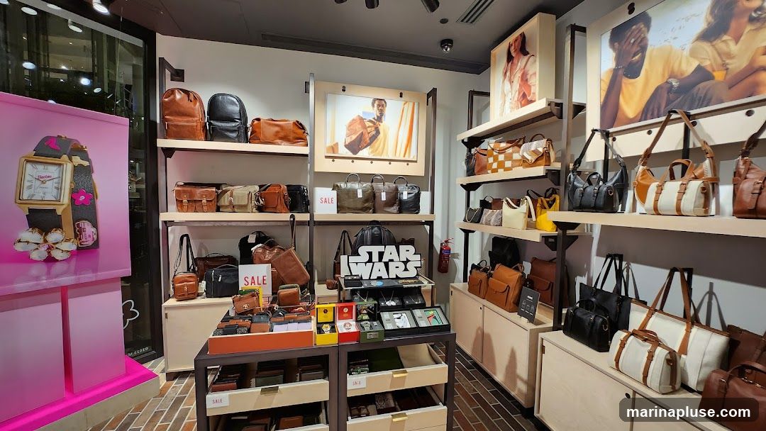 A well-lit interior view of a Fossil store, showcasing a variety of leather bags, wallets, and watches. The display features shelves stocked with different styles and colors of bags, from backpacks to crossbody bags. A prominent "Star Wars" themed display is visible, along with promotional images of models wearing Fossil products. Several "SALE" signs are placed throughout the store. On the left, a large advertisement for a Barbie-themed watch is displayed.