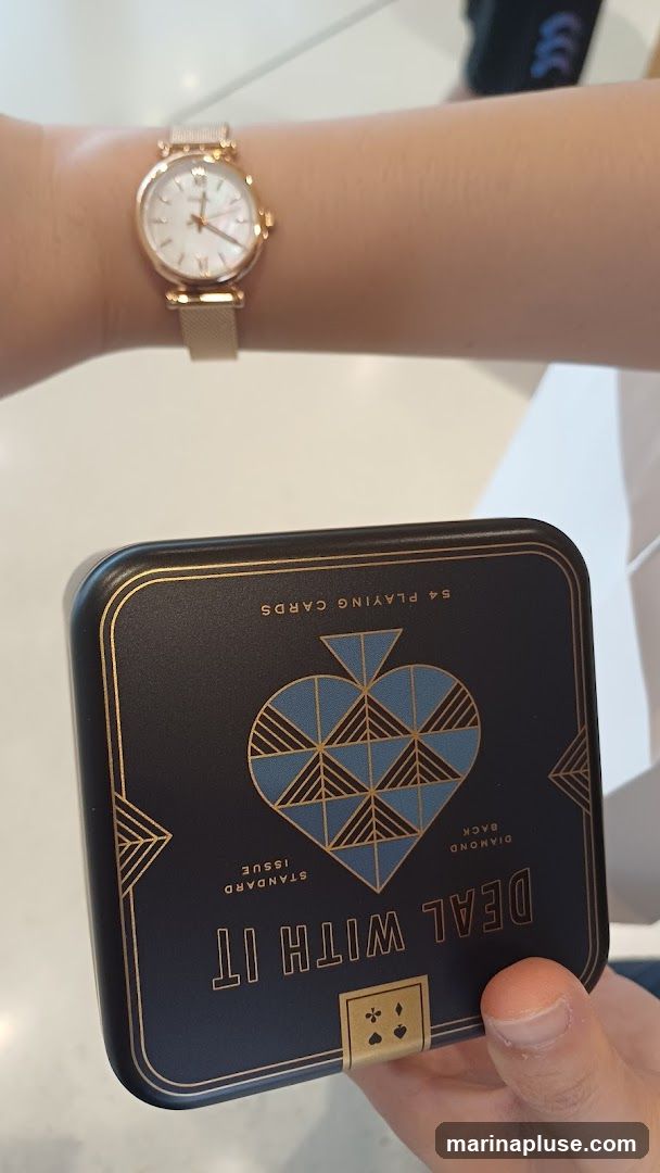 A close-up shot shows a person's wrist wearing a rose gold watch with a white face and Roman numerals. In front of the watch, a dark blue tin box with gold accents is held by a hand. The tin is decorated with a large spade symbol and text that reads "DEAL WITH IT STANDARD ISSUE DIAMOND BACK 54 PLAYING CARDS".