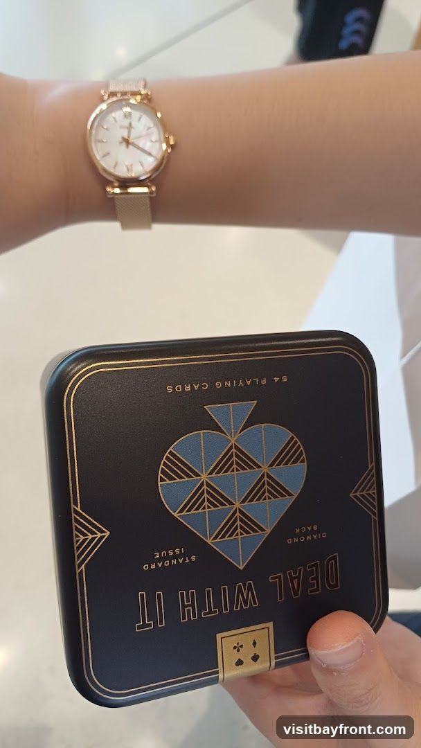 A close-up shot shows a person's wrist wearing a rose gold watch with a white face and Roman numerals. In front of the watch, a dark blue tin box with gold accents is held by a hand. The tin is decorated with a large spade symbol and text that reads "DEAL WITH IT STANDARD ISSUE DIAMOND BACK 54 PLAYING CARDS".