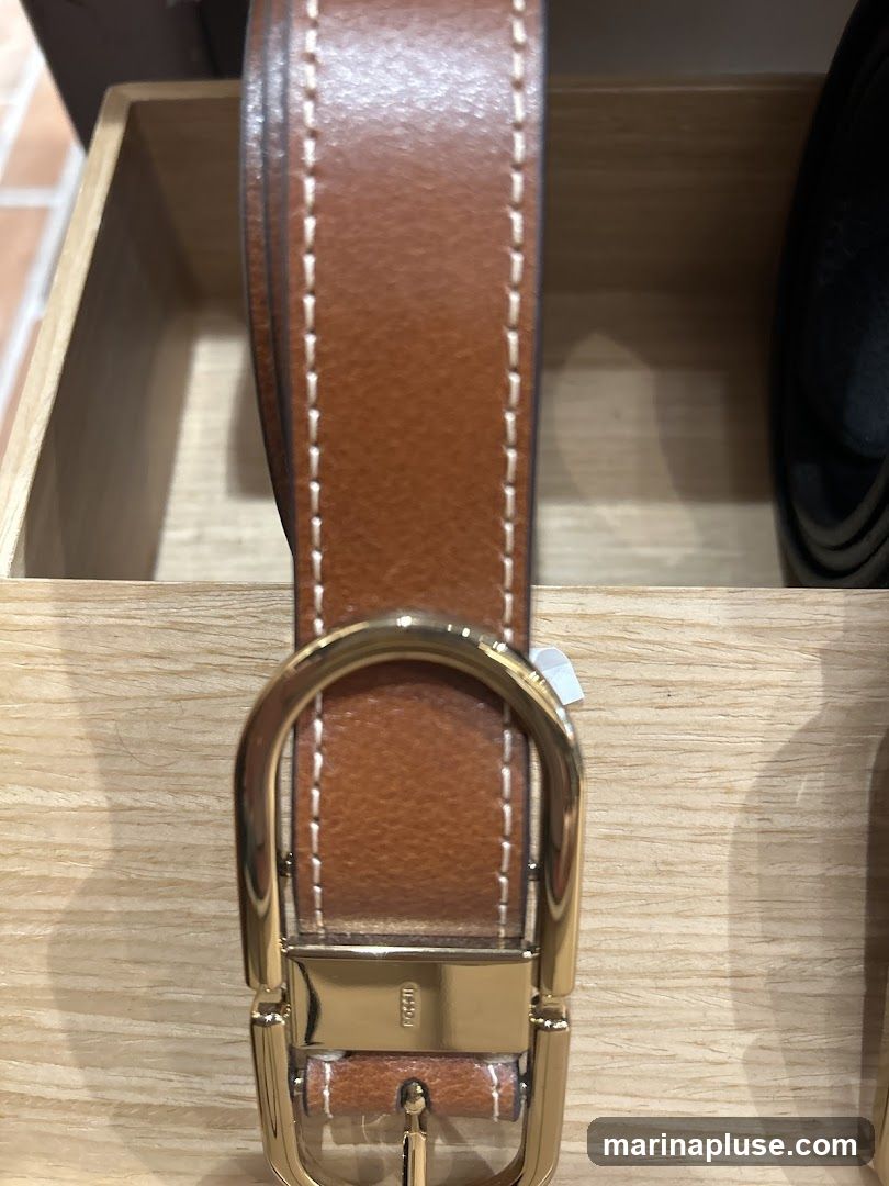 A close-up shot of a brown leather belt with white stitching and a gold buckle. The belt is displayed in a wooden box. The buckle has the word "FOSSIL" embossed on it.