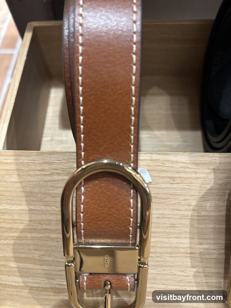 A close-up shot of a brown leather belt with white stitching and a gold buckle. The belt is displayed in a wooden box. The buckle has the word "FOSSIL" embossed on it.