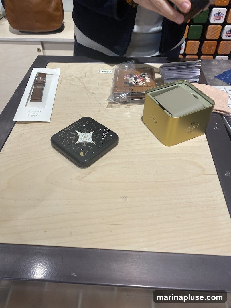 A wooden counter displays various Fossil brand items, including a watch strap in packaging, a wallet featuring Mickey and Minnie Mouse, and a gold-colored watch tin. A decorative tin with a celestial design is also visible. In the background, a person is handling an item, and a wall is decorated with colorful square tiles.