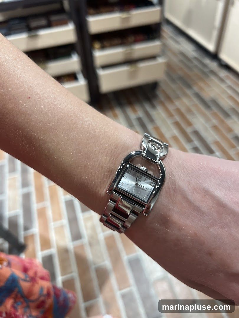 A close-up shot of a woman's arm wearing a silver watch with a rectangular face. The watch has a metal band and a silver dial with silver hour markers and hands. The background shows a tiled floor and shelves with various items.