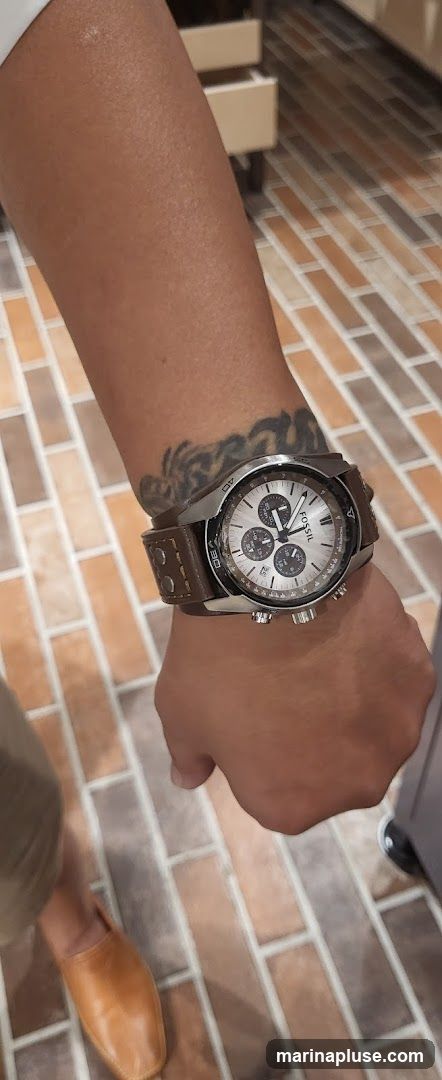 A close-up shot of a person's wrist wearing a Fossil watch. The watch has a silver dial with three subdials and a brown leather strap. The person has a tattoo on their forearm. The background shows a tiled floor and some furniture.