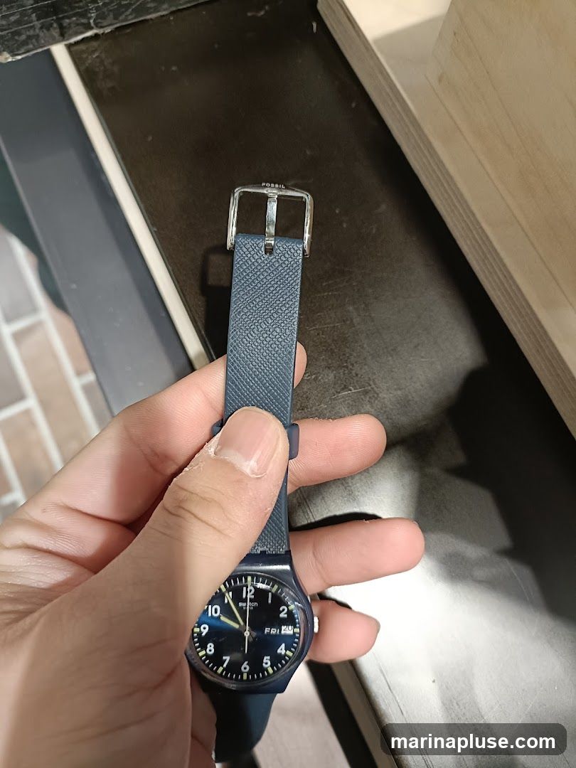 A hand holds a dark blue Swatch watch with a textured strap. The watch face is dark blue with white numbers and hands. The date window shows "FRI 24". The buckle of the strap has the word "FOSSIL" engraved on it.