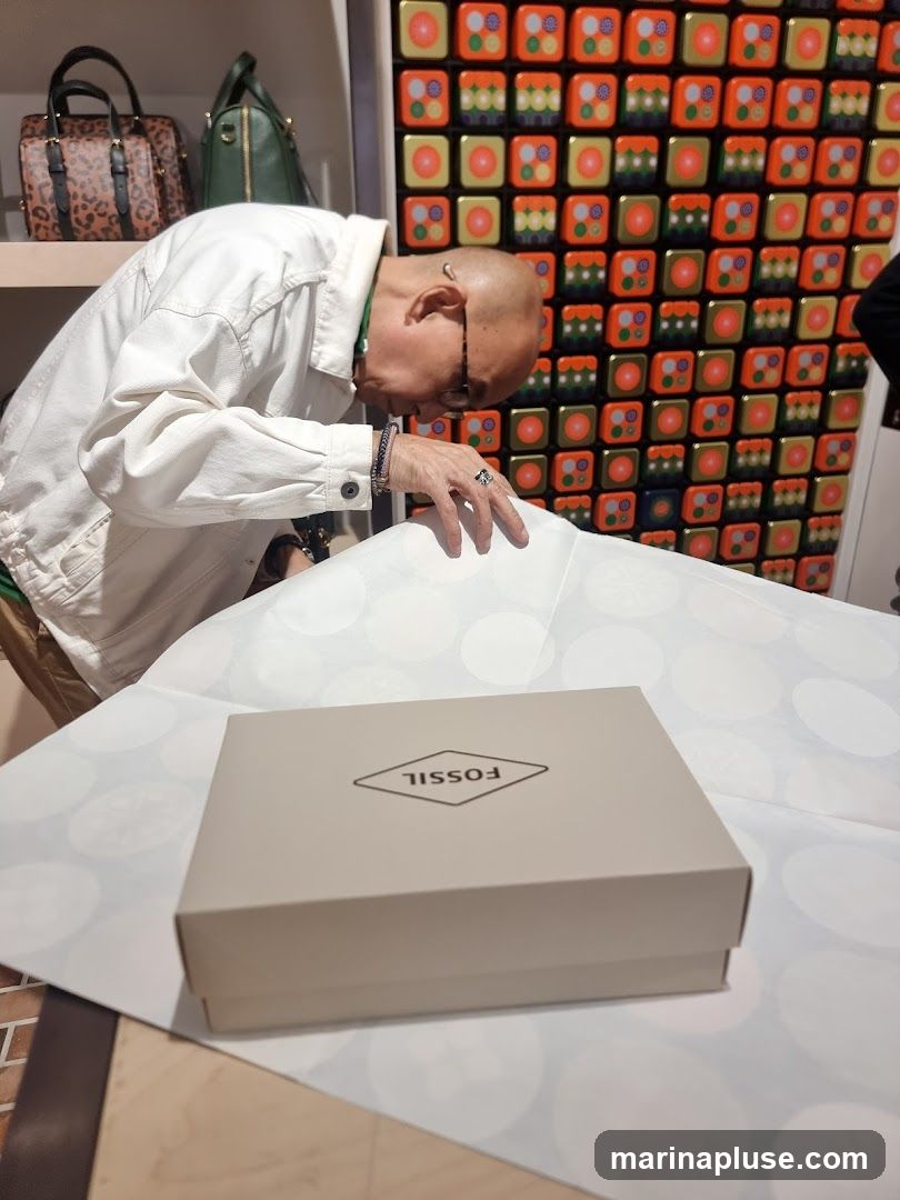 A person in a white jacket is leaning over a table with a Fossil branded box on it. Behind them, on a shelf, are two handbags, one with a leopard print and the other green. The wall behind is decorated with a grid of colorful square tiles.