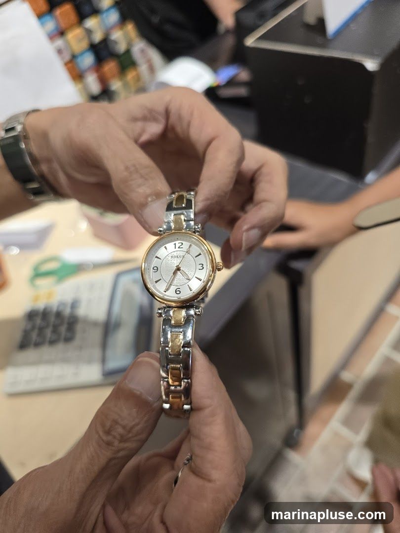 A person is holding a Fossil watch with a silver and gold band and a white face. The watch has Roman numerals and a small second hand dial. In the background, there is a calculator and other items on a counter.