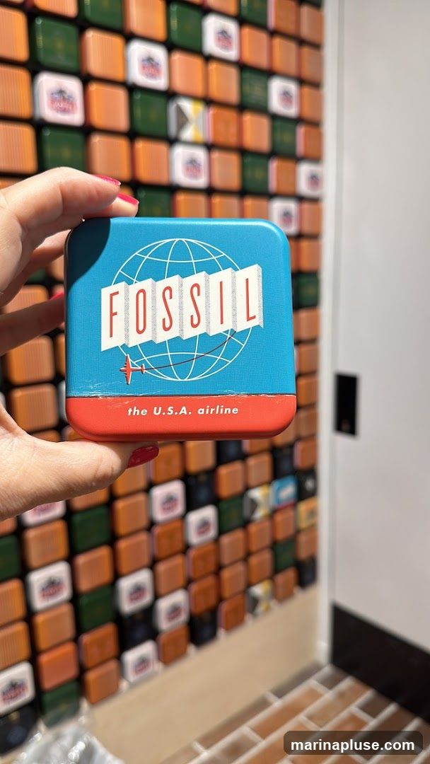A hand holds a blue and red tin with the Fossil logo, featuring a globe and an airplane. The tin reads "FOSSIL" and "the U.S.A. airline". The background is a wall covered in small, square tiles in various colors, including orange, green, and white, some with a red logo.