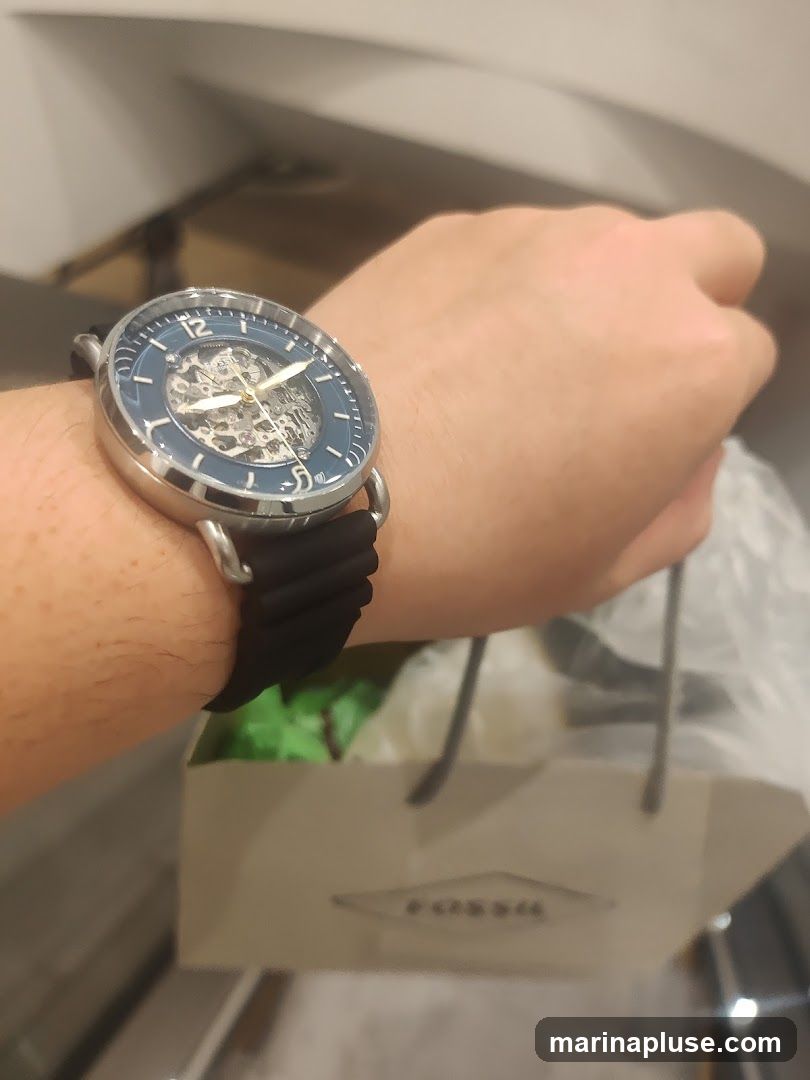 A person's wrist wearing a skeleton watch with a blue bezel and black strap. In the background, a light-colored shopping bag with the Fossil logo is visible.