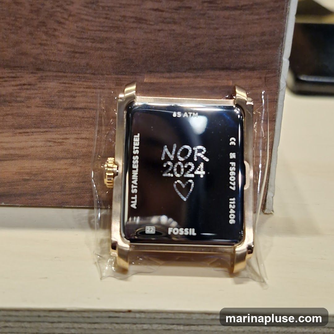 A close-up shot of the back of a Fossil watch. The watch case is rose gold colored and the back is black with engraved text. The engraving reads "NOR 2024" with a heart symbol below it. Text around the edges indicates "ALL STAINLESS STEEL" and "65 ATM". There are also some certification marks and a model number "FS6077 112406". The Fossil logo is visible at the bottom. The watch appears to be new and is still in its protective plastic packaging. The background is a textured wooden surface.