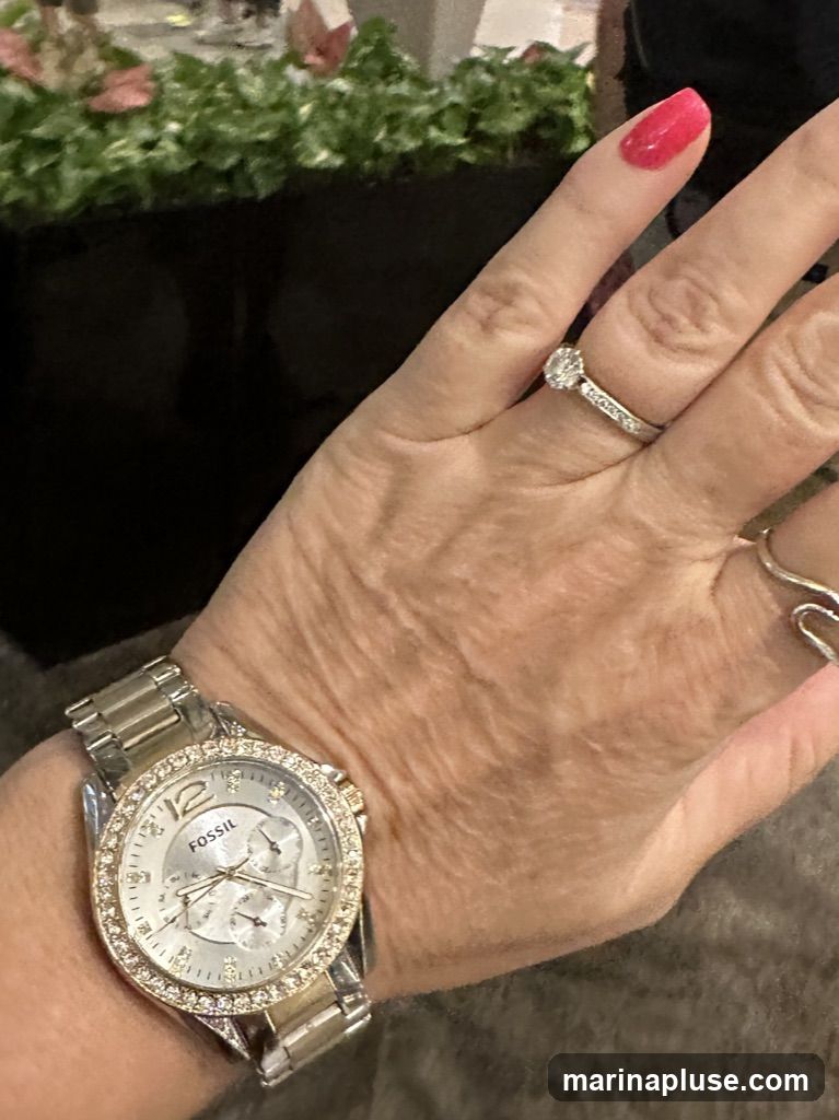 A close-up shot of a woman's left hand, adorned with a silver Fossil watch featuring a diamond-encrusted bezel and a silver band. She is also wearing a diamond engagement ring and a coiled silver ring on her right hand. Her fingernails are painted a bright pink. In the background, out of focus, are green plants and a dark surface.