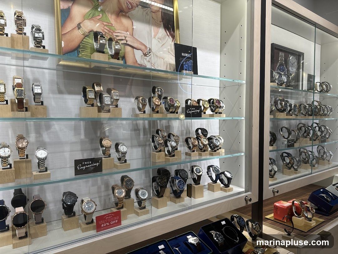 A display case filled with various watches. The watches are arranged on shelves and stands, with some watches in boxes. There is also a promotional sign for free engraving and a sign for select watches 40% off. A large advertisement featuring two women wearing watches is displayed at the top of the case.