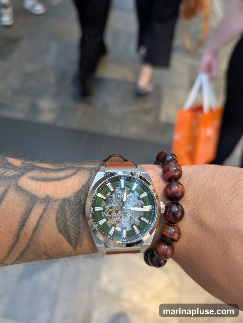 A close-up shot of a person's wrist wearing a silver Fossil watch with a green skeleton dial and a brown leather strap. A beaded bracelet made of reddish-brown stones is also visible on the wrist. In the blurred background, people are walking, and one person is carrying an orange shopping bag.