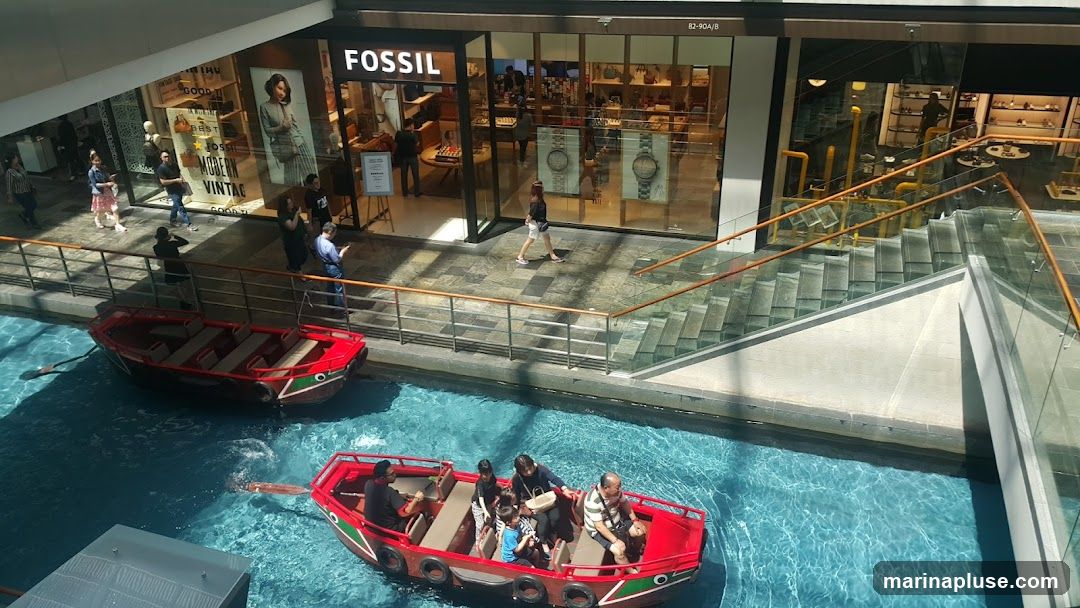 The exterior of a Fossil store with its prominent logo above the entrance. Inside, shelves display watches and accessories. People are seen walking past the store and inside. In the foreground, red boats with passengers navigate a canal.